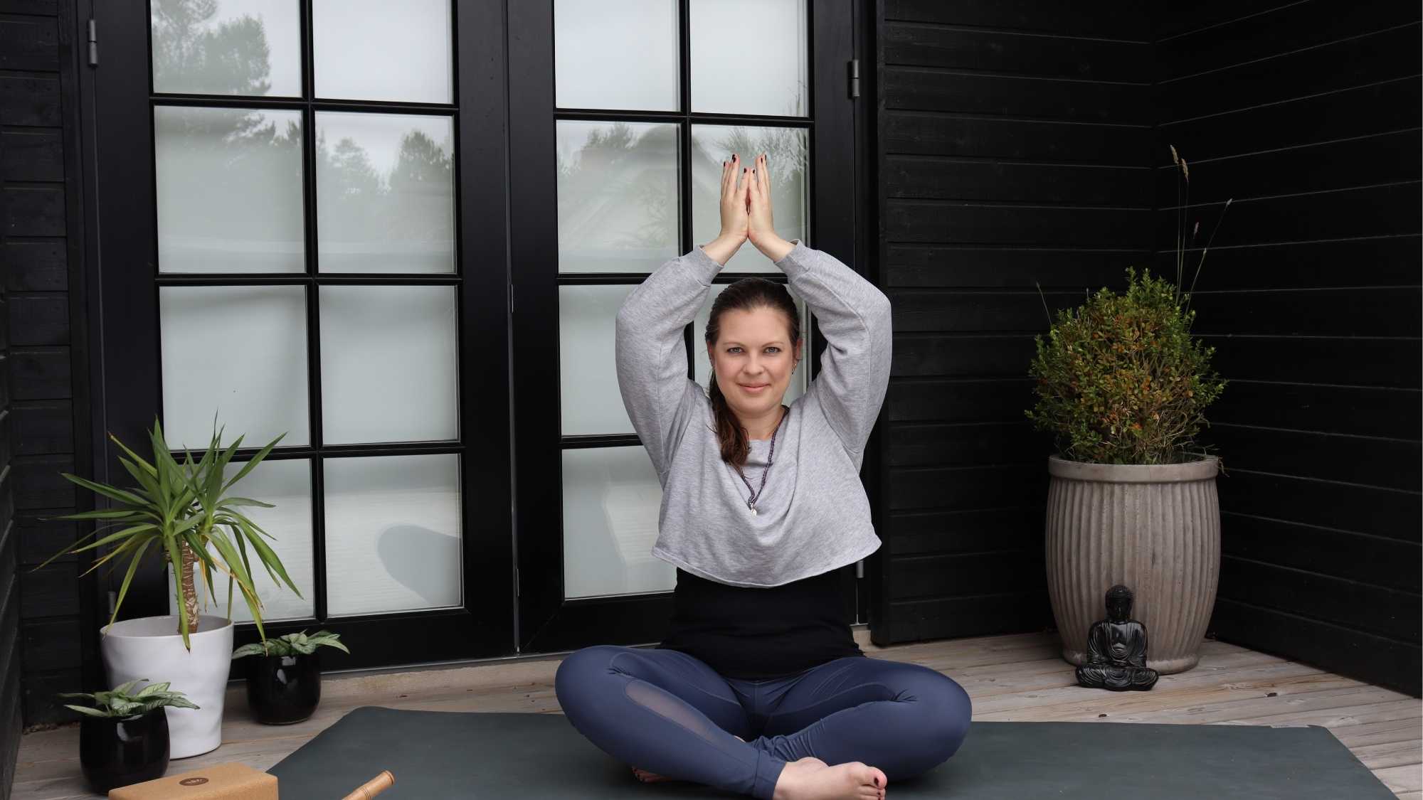 Eine Frau sitzt auf einer Yogamatte. Sie hält die Hände über dem Kopf zu einer Lotusblüte geformt.